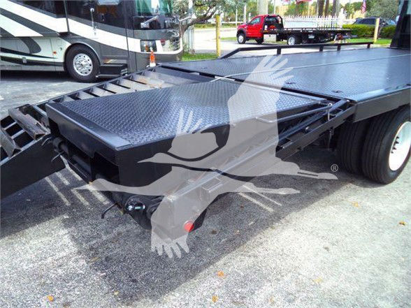 Flatbed truck trailer with diamond plate deck and hydraulic pivoting ramps; Freightliner M2 chassis visible in background.