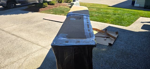 Long rectangular Volvo truck cab component or part in a driveway, taped cardboard, and packaging visible; light brown protective wrap and black plastic base.