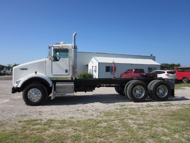 Kenworth T800 truck chassis with cab in foreground, white cab, chrome exhaust stack, long wheelbase with dual rear axles and empty flatbed area.