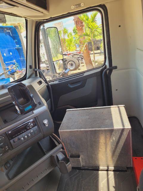 Volvo VNR daycab interior showing dashboard with gear selector, steering area, and a rectangular metal box on the floor beside the passenger seat; window view of palm trees outside.