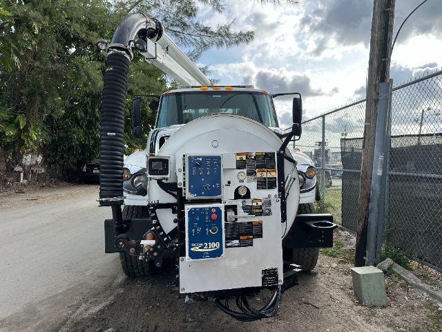 Back of a white vacuum truck with a circular rear tank and blue control panels. Visible labels and warning stickers on the panel doors, hoses, and wiring harness.