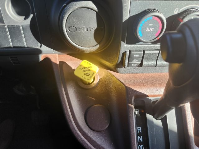 Hino truck cab interior showing steering wheel, A/C control, yellow parking brake handle cap reading 'PARKING BRAKE', and gear selector with PRND.