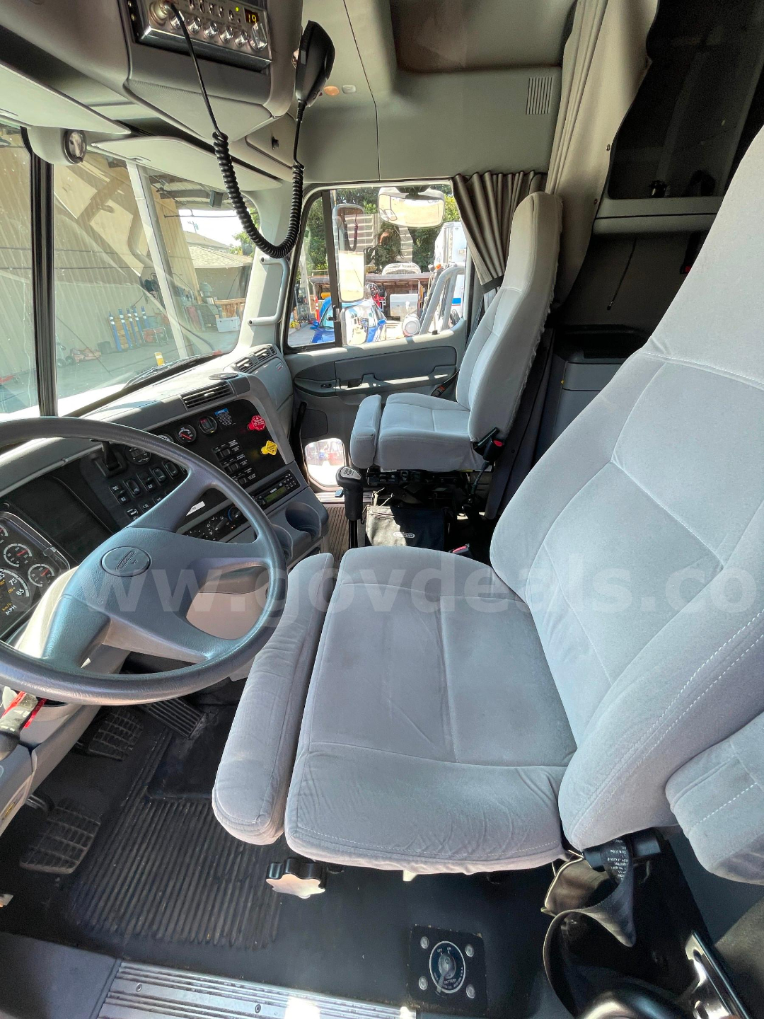 Cab interior of a 2004 Freightliner Columbia 120 showing steering wheel, dashboard with gauges, driver seat, and overhead console with microphone; light gray cloth seating.
