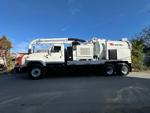 Large white vacuum truck with hose reel in front and mounted vacuum unit, side view showing ladder and equipment; Vactor/VacCon branding on the side tank.