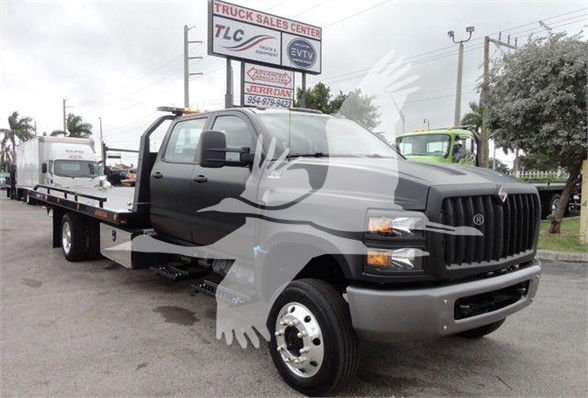 Tow truck with flatbed in a dealership lot. Visible text includes a TLC Truck Sales Center sign in background and a J98978 stock number. The truck is a new 2023 International CV with a dark gray front grille, large bumper, dual rear wheels,