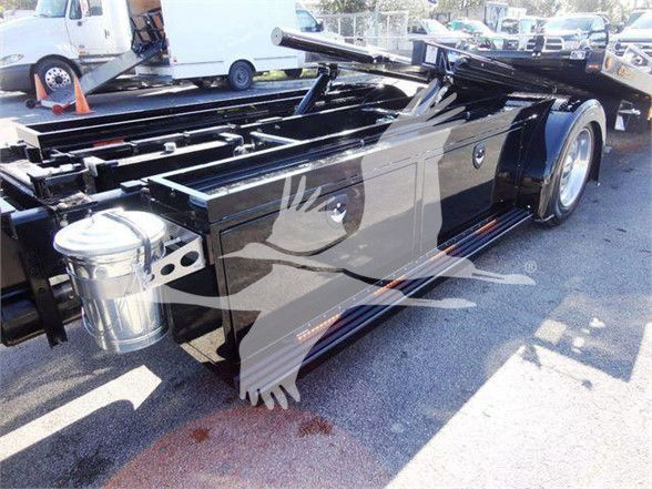 Truck chassis with a long wheelbase and a polished metal fuel tank. Visible components include a glossy black frame, chrome exhaust/muffler, and wheel with heavy-duty tire. There is a watermark overlay of a bird graphic on the image.
