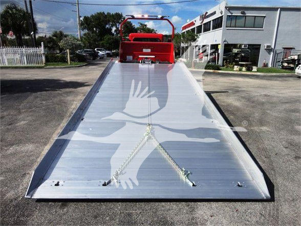 Tow truck bed with aluminum platform, hydraulic rear ramps, and red rollover protection visible at top; bright dealership building in background.