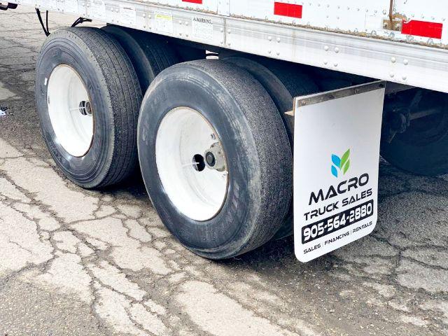 Trailer rear view showing dual rear axles with black tires and white rims, plus a vertical white mudflap displaying MACRO TRUCK SALES logo and contact number.