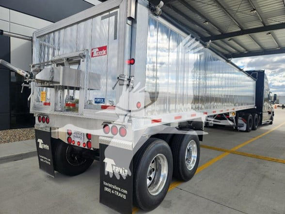 Travis dump trailer in a parking lot showing dual rear axles, polished aluminum sides, rear gate, and tires with mudflaps displaying Travis logo.