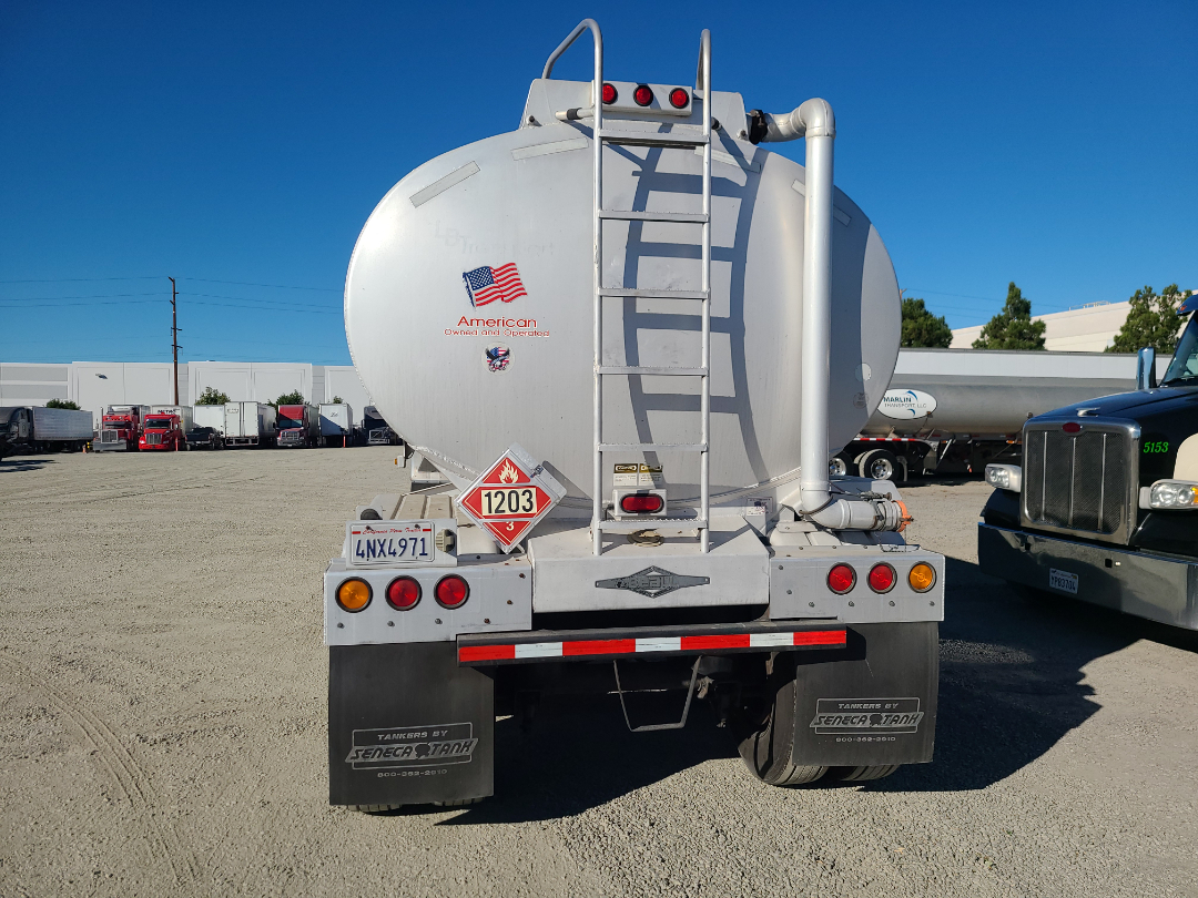 Back view of a 1998 Beall semi-trailer with a circular tanker, ladder on the back, American sticker, hazard placard 1203, and multiple tail lights.