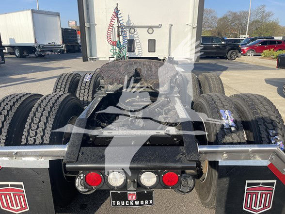 Rear view of a Kenworth truck chassis with fifth wheel and multiple wheels visible; trucks and trailers in background.