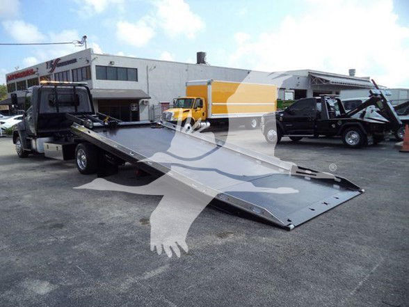 Tow truck with flatbed extended, 2014 Hino 258ALP in lot, other vehicles in background.