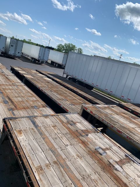 Flatbed trailer deck with wooden planks and side railing, long cargo area, blue sky with clouds in background.