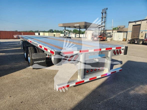 Flatbed trailer by Wabash, dual-axle setup, rear lights and reflective tape visible, long aluminum bed, storage toolbox at side, industrial lot background.