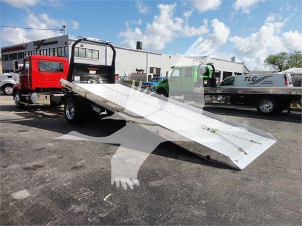 Truck with a tilted aluminum tow bed/ ramps in a parking lot. Visible text on the truck bed and nearby equipment not legible.