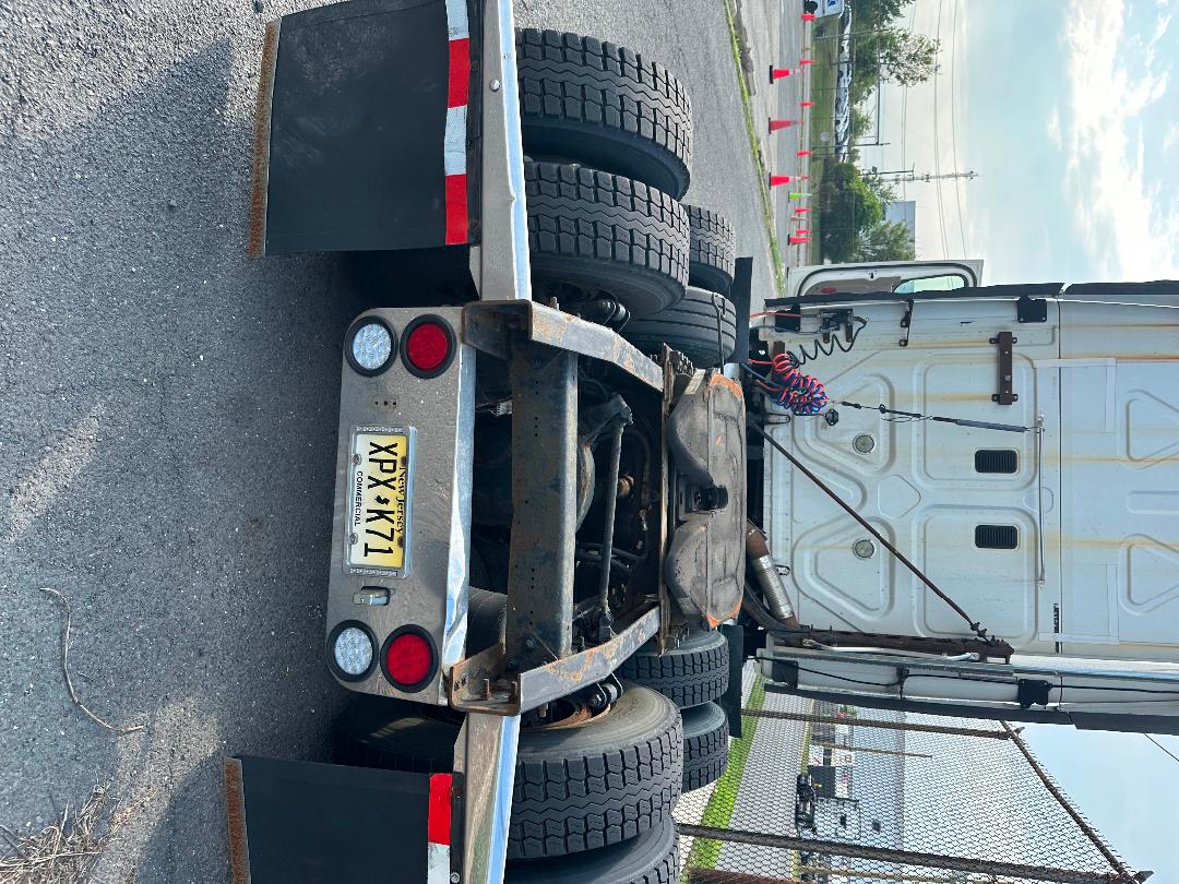 Flatbed trailer with dual rear axles and exposed drivetrain showing spare wheel mounts and a yellow license plate YP... visible text on plate and truck rear.
