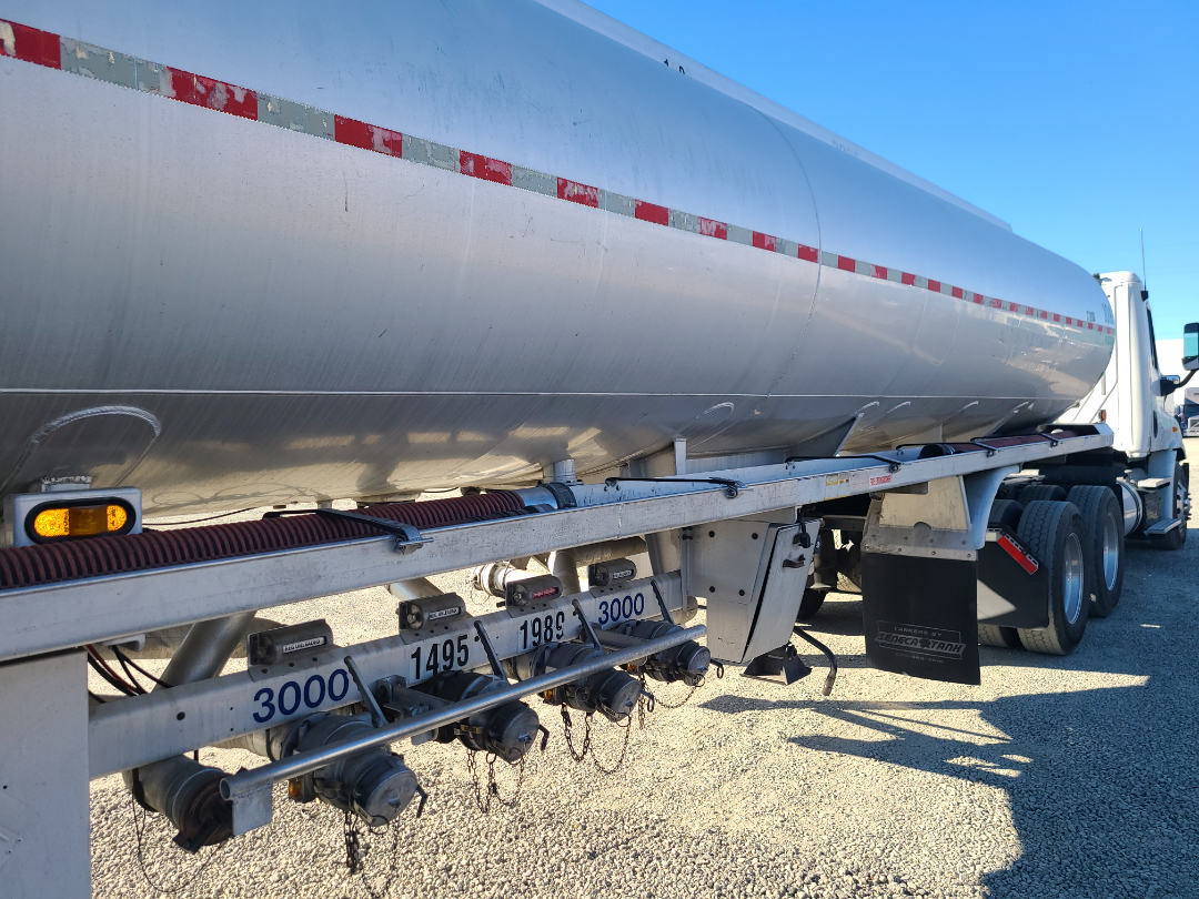 Beall semi trailer with large cylindrical aluminum tank showing inspection ports, hoses, and valve controls along frame; multiple numbers and a red/white reflective stripe on the tank.