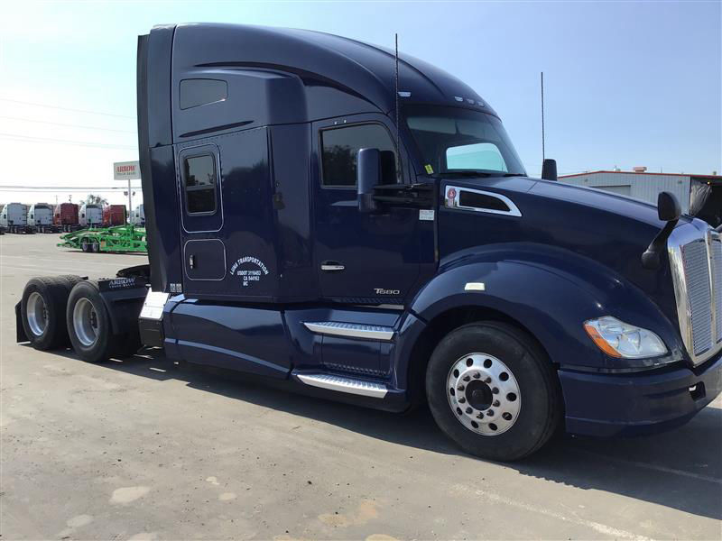 Blue 2016 Kenworth T680 semi-truck in a parking lot with sleeper cab and chrome steps. Side shows door and window, with wheels and dual rear axles.