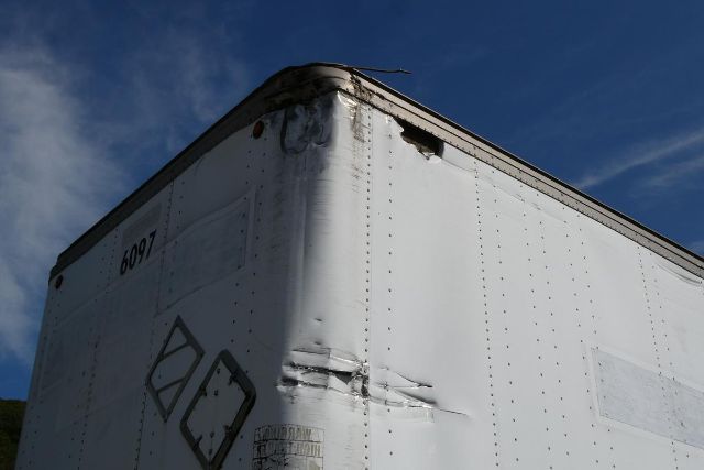 Vehicle trailer corner with riveted metal surface, number '6097' on door and damaged corner area.