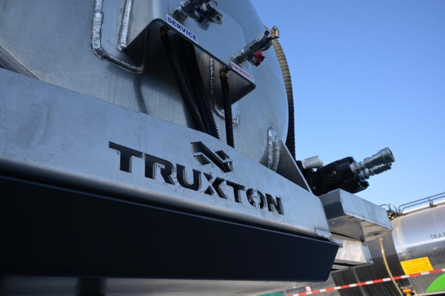 Truxton vacuum trailer with aluminum exterior showing 'TRUXTON' branding and hoses/components near top with a blue sky.