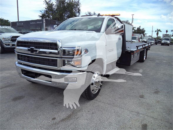 Chevrolet Silverado 6500HD tow truck with flatbed, front grille with Chevrolet emblem, white cab, amber light bar on roof, side mirrors, and extended flatbed. Visible text includes Chevrolet badge and details on the truck.
