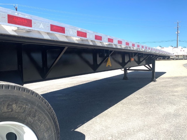 44- or 48-foot flatbed trailer undercarriage with black steel frame, side rails, reflective red and white markings, landing gear, and a wheel visible on left; blue sky.