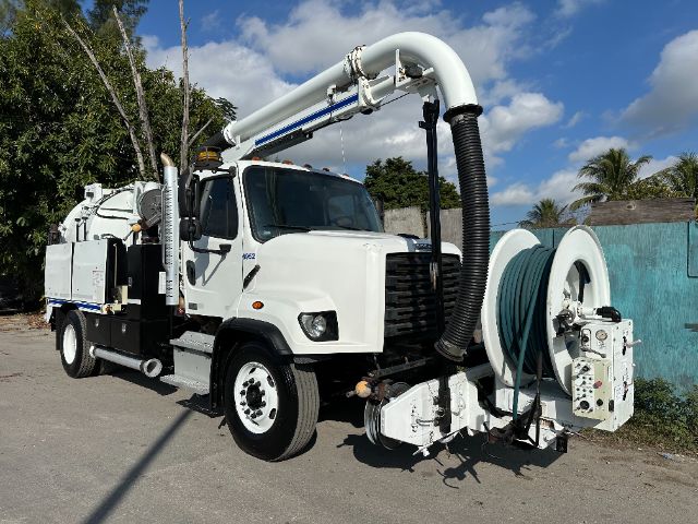Freightliner 108SD vacuum truck with large hose reel and boom in front, white cab and equipment body, blue hose coiled on reel, clear blue sky
