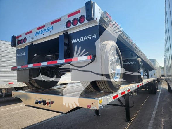 Wabash flatbed trailer showing rear with spare tire, taillights, and reflective tape. Brand name WABASH visible on sides and rear; trailer on a loading dock with black undercarriage.