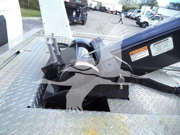 Tow truck platform with hydraulic winch and hook assembly on diamond plate deck. Visible labels, warning sticker, and a chrome/metal track.