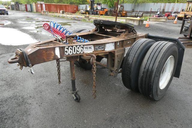 Trailer dolly with dual rear wheels and coiled air hoses; rusty frame and front hitch, tiedown chains, number plates, and steel post on ground.