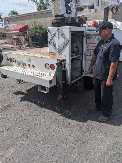 Truck-mounted aerial lift with control panel and stabilizing legs, man standing beside unit, rear view showing platform and safety decals.