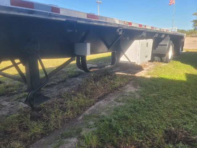 Flatbed trailer undercarriage showing landing gear, frame rails, and dual rear axles with tires. Side rails and reflectors visible along top edge.