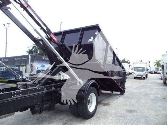 Truck bed with a black dump-style dump body, rear wheel visible, hydraulic arms extending to dump the bed, Freightliner-style refuse truck in a dealership lot.