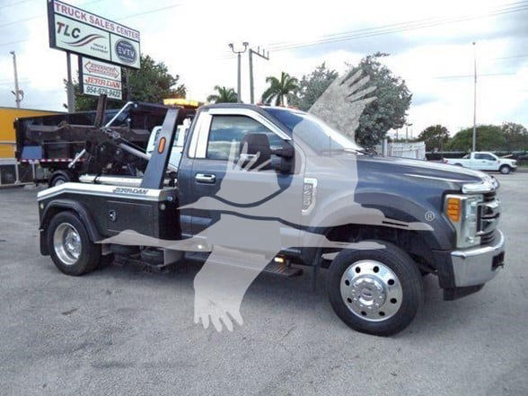 Ford F-450 tow truck with flatbed and wheel lift seen in a dealership lot; visible text includes brand details on the truck and background signs.