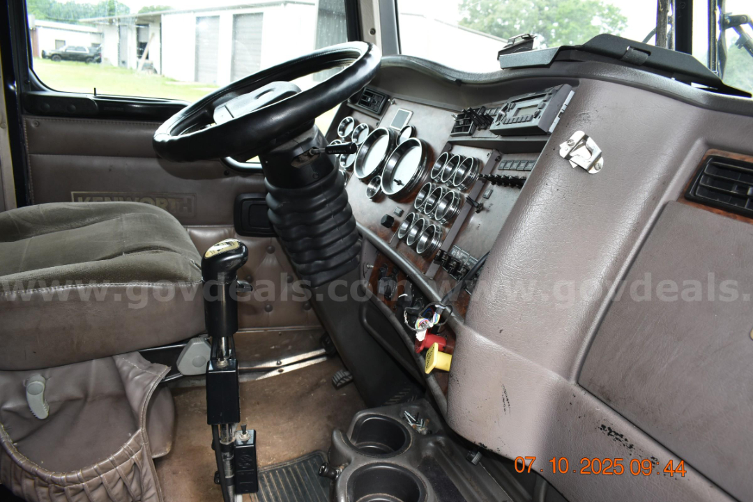 Kenworth W900 cabin interior showing steering wheel, dashboard with multiple gauges, gear selector, and control panel.