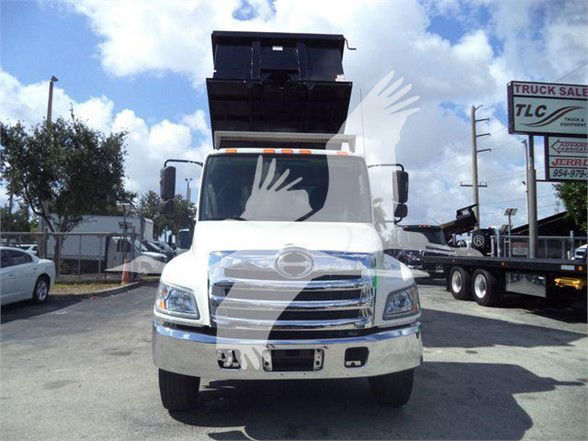 Front view of a white 2016 Hino 268A dump truck with a raised dump bed. Visible text includes Hino logo on grille and a sign in background; the truck has large chrome grille and bumper.