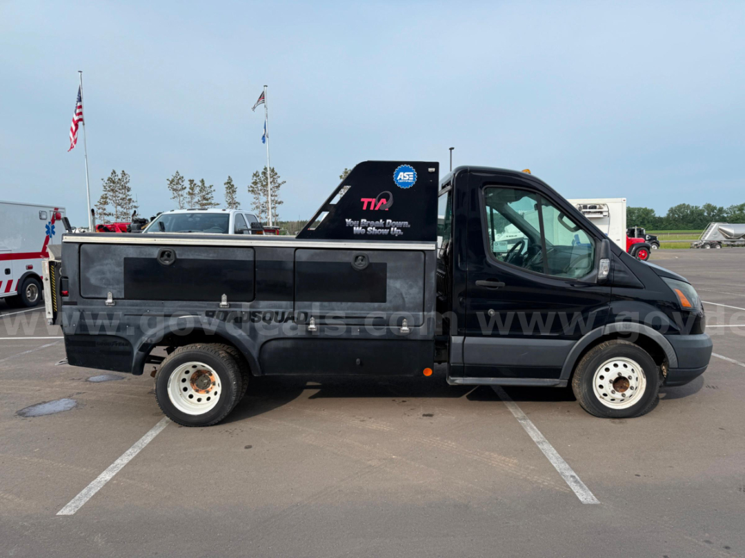 Ford Transit cargo/service utility truck with storage compartments along the bed and crew cab, black exterior, lettering on rear glass, roadside parking.