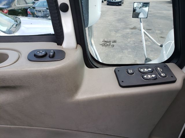 Vehicle interior window view with mirror and control panel. Text on window near mirror reads 'MACK TRUCK... INDIA' etc. The door panel shows circular window switch and side mirror control. Exterior shows partial reflection of other trucks.