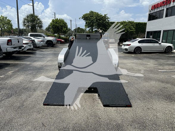 Flatbed trailer loading ramp with diamond plate, black ramp surface, white pickup trucks in lot. No visible part numbers or brands readable.