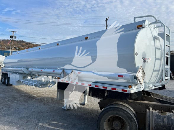 Truck trailer showing a white 9200-gallon trough bottom petro tank with rear ladder and support legs, side piping, and wheel hub visible.