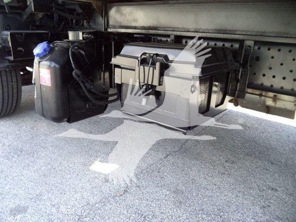 Underbody view of a box truck showing a portable toolbox and a coolant reservoir with a blue cap. Visible text on the reservoir label and toolbox not legible.