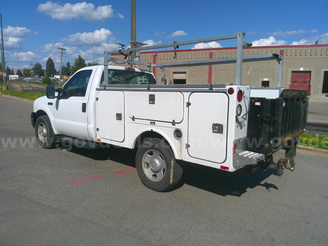 White Ford F-350 utility service truck with multiple tool compartments, ladder rack, and stake bed viewed from side-rear angle.