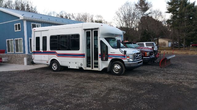 2009 Ford E-450 shuttle bus photographed in a lot with a plow truck attached to its front; white body with blue/red stripe, multiple windows, service doors, and a blue building in background.