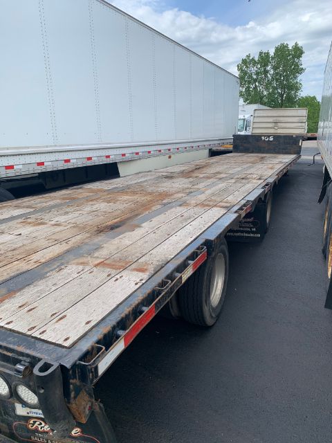 Flatbed trailer side with weathered wooden deck, Doonan logo on mudflap, steel frame, rear axle, and wheel. Weathered boards show wear.