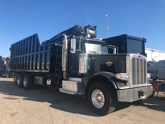 Peterbilt 388 heavy-duty dump truck with grapple, dark green cab, chrome grille, large side exhaust, and crane on top, parked outdoors