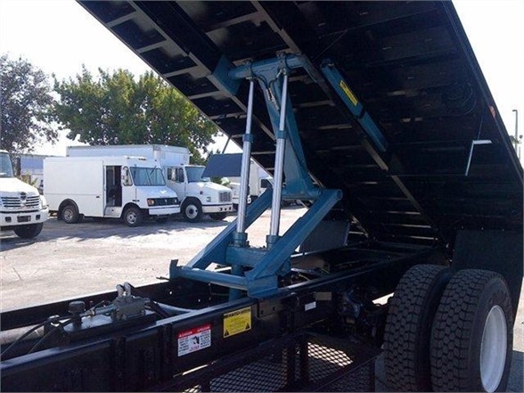 Truck flatbed body with hydraulic raise and tilt system in up position; blue mechanism and support arms visible; rear dual wheels visible; stock area in background.