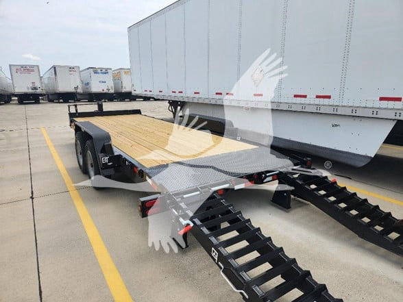 Flatbed trailer with dual fold-out ramps and wooden deck, parked in a lot next to a large white trailer.