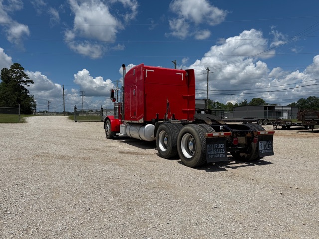 Red 2007 Peterbilt 379 semi truck with sleeper cab, rear view showing dual rear axles and mud flaps with white text, on a gravel lot under a blue sky with clouds.