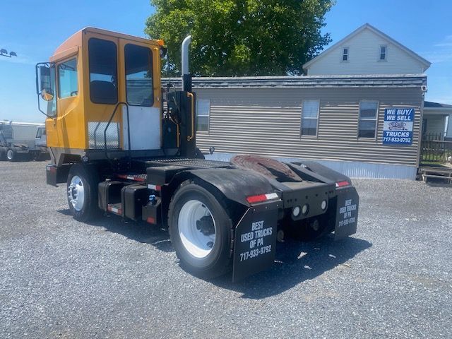 Truck tractor chassis with yellow cab on a gravel lot, rear view shows fifth wheel and white wheels.