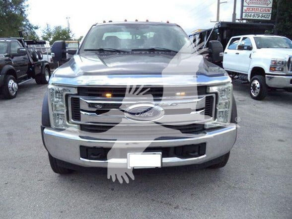 Ford truck front view showing grille with Ford emblem, headlights, and front bumper. Plate area blurred. Watermark overlay of a handshake graphic across the image.